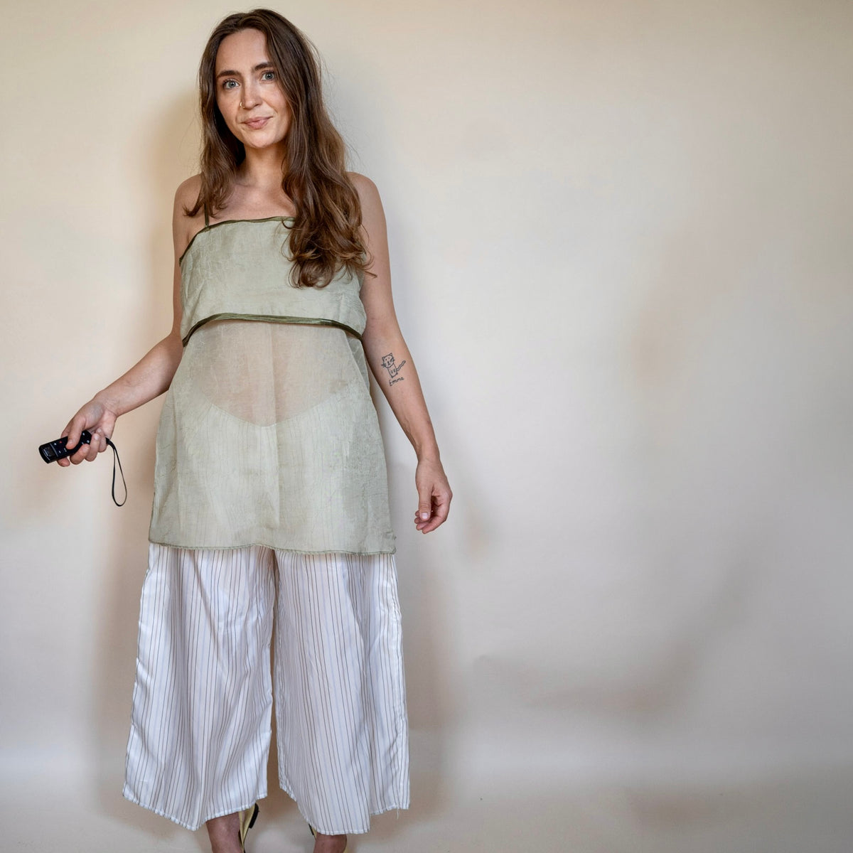 A woman modeling a sleeveless sage green sheer mesh top with a solid waistband, paired with white and gray vertically striped cropped pants, standing against a plain beige wall.