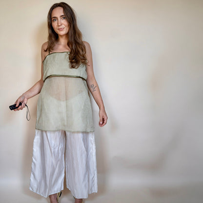 A woman modeling a sleeveless sage green sheer mesh top with a solid waistband, paired with white and gray vertically striped cropped pants, standing against a plain beige wall.