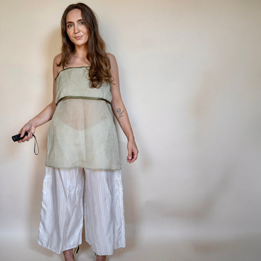 A woman modeling a sleeveless sage green sheer mesh top with a solid waistband, paired with white and gray vertically striped cropped pants, standing against a plain beige wall.